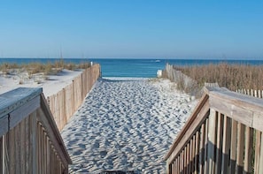 Ein Holzsteg führt zu einem Strand mit Sand und spärlicher Vegetation.