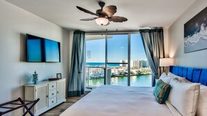 A hotel room with a large bed, a ceiling fan, a TV, a view of the ocean, and a balcony with curtains. - Luxurious Condo with Beach Access and Ocean Views (Destin)