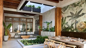 An open-plan dining area with a long table set for six, surrounded by wooden chairs. There is a small garden area with plants and a bench. The space has a high ceiling with wooden beams and a large window providing natural light. - Casa San Antonio, Designer Villa w / Rooftop Pool. (Cartagena)