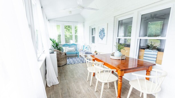 Un interior luminoso y blanco con una mesa de comedor de madera y sillas blancas. Hay ventanas con cortinas, un ventilador de techo y vistas a zonas verdes al exterior.