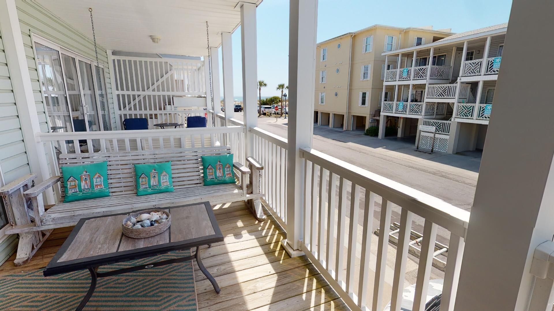 A wooden porch with a table and chairs, a bench with cushions, and a view of a building across the street.