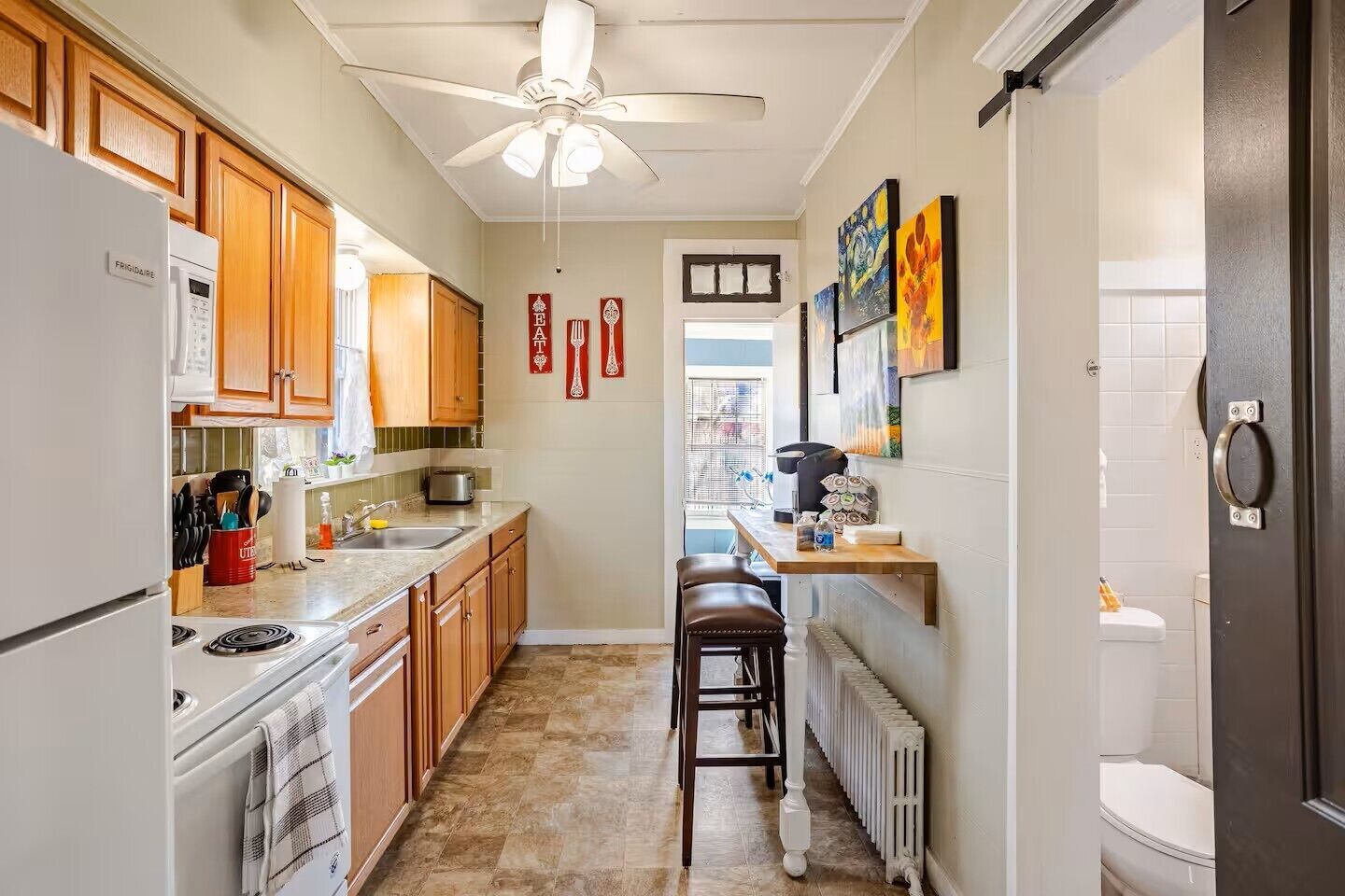 A compact kitchen with wooden cabinets, a built-in oven, a refrigerator, and a ceiling fan. There are stools at the counter and a radiator on the floor.