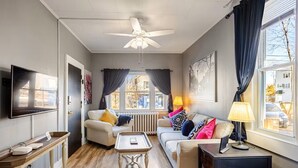A cozy living room with a ceiling fan, a TV mounted on the wall, a sofa with colorful cushions, a coffee table, and a lamp.