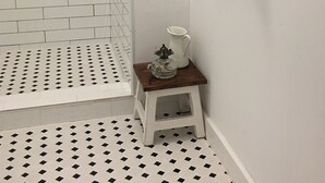 Een badkamer met betegelde vloer, een douchegedeelte, en een klein houten krukje met een waterkan en glas erop.