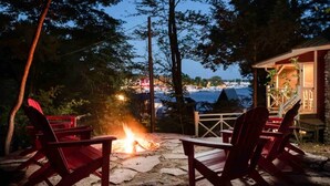Une terrasse en pierre avec deux fauteuils Adirondack donnant sur un foyer extérieur, et offrant une vue panoramique sur une étendue d’eau et des bâtiments au loin.