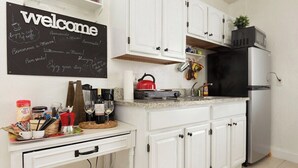 A kitchen with white cabinets, a black refrigerator, a chalkboard with 'welcome' and various greetings, and a coffee station with cups and a kettle. - 1 Bedroom Apt near Coconut Grove (Miami)