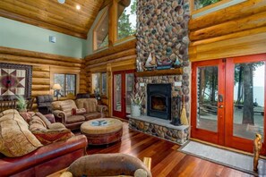 L’intérieur d’une cabane en rondins, avec une cheminée en pierre, un plancher en bois et une grande fenêtre donnant sur un paysage naturel.