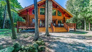 A log cabin with a porch, a stone wall, and a gravel driveway surrounded by trees. - Fully Loaded Lakefront Lodge w/ Pier & Game Room (Sturgeon Bay)