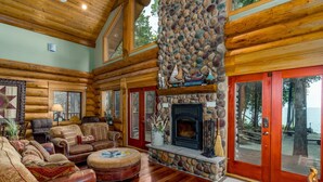 A log cabin interior with a stone fireplace, wooden furniture, and a view of the outdoors. - Fully Loaded Lakefront Lodge w/ Pier & Game Room (Sturgeon Bay)
