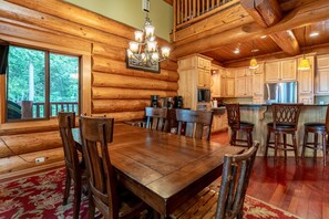 A log cabin interior with a dining area, kitchen, and a large window overlooking a forest.