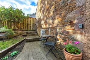Una terraza de madera con una mesa y sillas, una planta en maceta con flores y un muro de piedra.