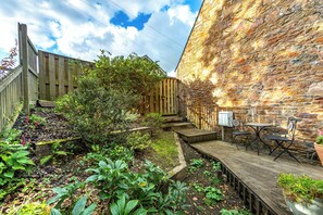 Una terraza de madera con un muro de piedra, una valla de madera y un pequeño jardín con plantas y un banco.