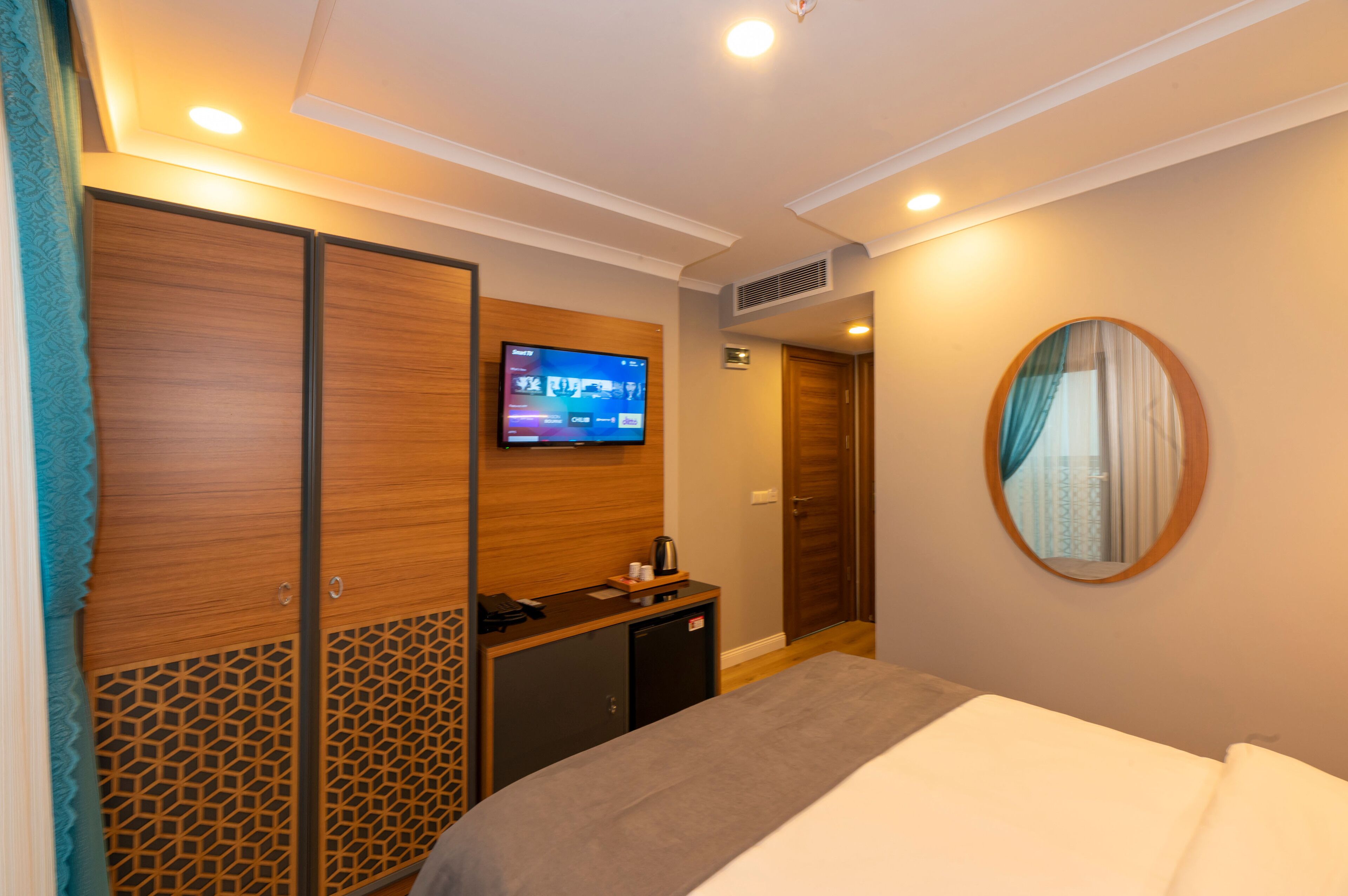 A hotel room with a bed, a TV mounted on the wall, a small desk with a kettle, and a wardrobe with a geometric pattern.