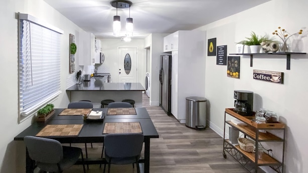Une cuisine moderne dotée d’un îlot central, d’un coin repas avec une table et des chaises, et d’un espace dédié au café, avec une étagère et une poubelle.