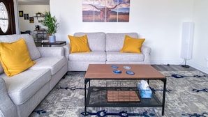 Un salon avec un canapé gris, deux coussins de couleur jaune moutarde, une table basse ornée d’assiettes décoratives et un tableau accroché au mur.