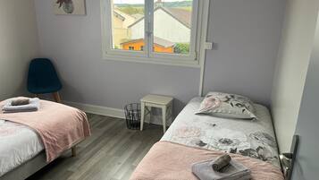 A bedroom with two beds, a window with a view of buildings, a chair, and a small table.