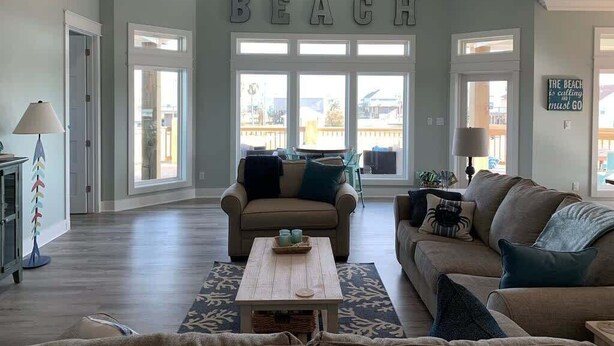 A spacious living room with a large window, a ceiling fan, and a 'BEACH' sign on the wall. - St. Somewhere, Dog Friendly (Crystal Beach)