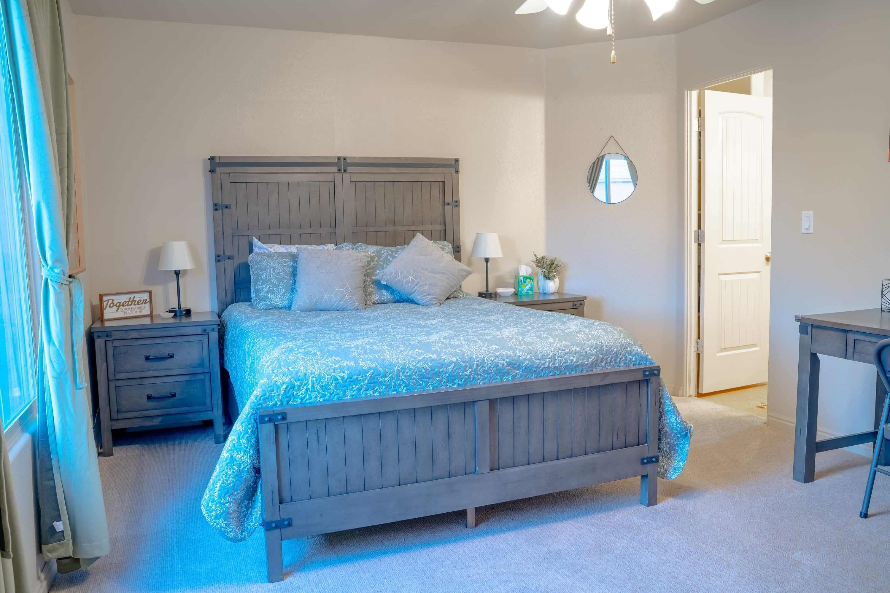 Een slaapkamer met een houten bed, twee nachtkastjes met lampen, een klein nachtkastje met een plant, en een deur naar een andere kamer.