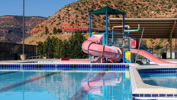 A swimming pool with a water slide and a covered area, surrounded by a fence and trees, with a hilly landscape in the background. - Spacious Townhome in Kanab! Near Zion, Bryce, and more! Pool and Hot Tub. Sleeps 8! (Kanab)