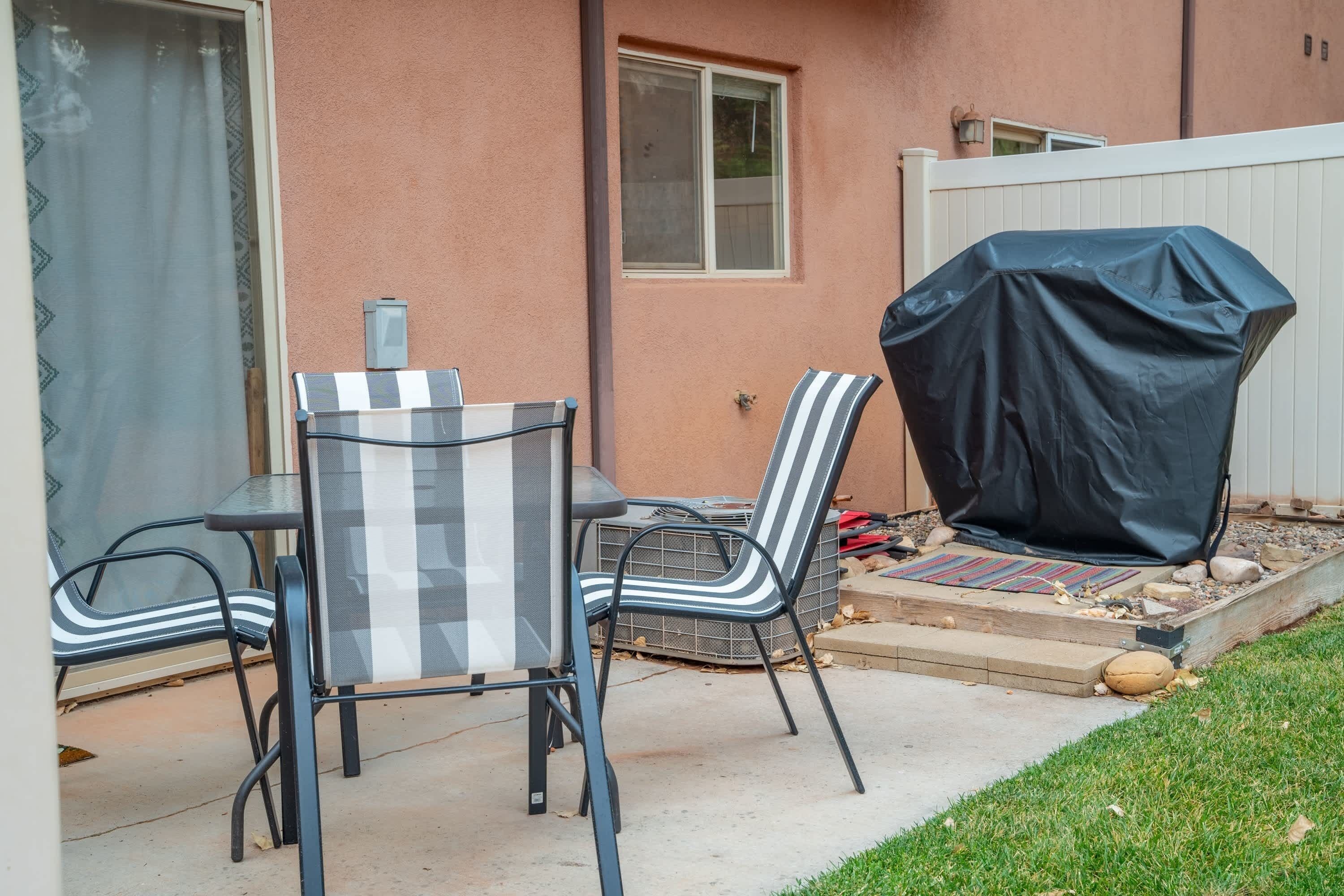 An outdoor patio with a table and chairs, a covered grill, and a white fence.