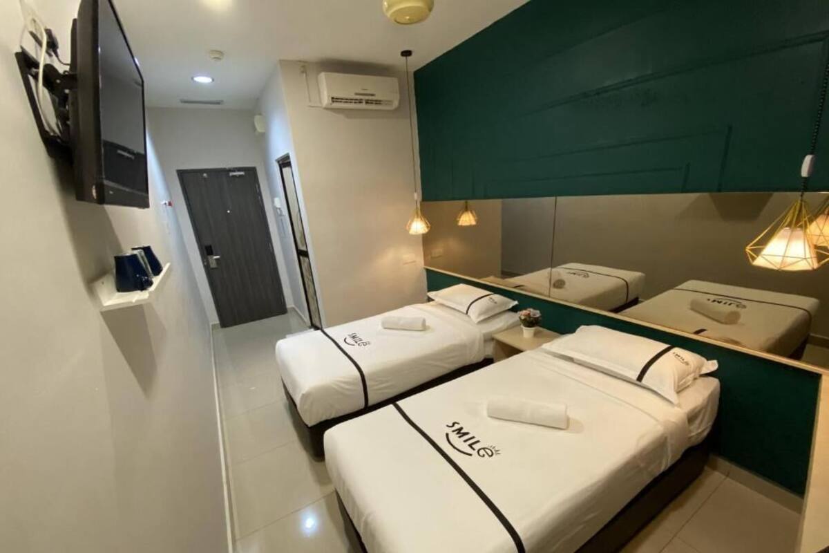 A hotel room with two beds, each with a white mattress and matching pillows, a flat-screen TV mounted on the wall, a small shelf with a blue object, and a mirror reflecting the room's interior.