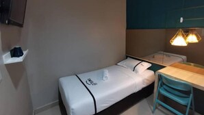 Een kleine kamer met een enkel bed, een bureau en een stoel, een tv aan de muur en een plank met een koffiezetapparaat.