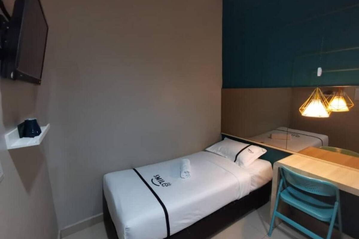 A small, single-bed room with a desk and chair, a wall-mounted TV, and a shelf with a coffee maker.