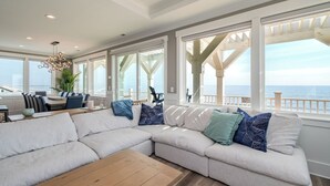 Uma sala de estar moderna com um sofá seccional grande, uma mesa de centro de madeira e uma área de jantar com mesa e cadeiras. Janelas amplas oferecem vista para o oceano.