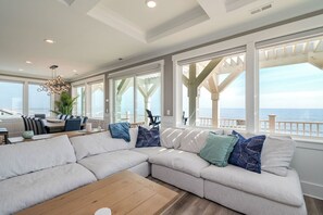 A modern living room with a large sectional sofa, a wooden coffee table, and a dining area with a table and chairs. Large windows offer a view of the ocean. - Saltwater Blessing: Beautiful Oceanfront Home with Private Pool, Elevator and Gr (Oak Island)