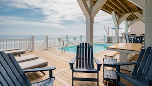 Um deck de madeira com espreguiçadeiras e uma mesa, com vista para uma piscina e o oceano.