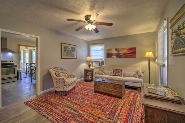 A living room with a ceiling fan, a sofa, a patterned armchair, a coffee table, a colorful rug, and a bookshelf.
