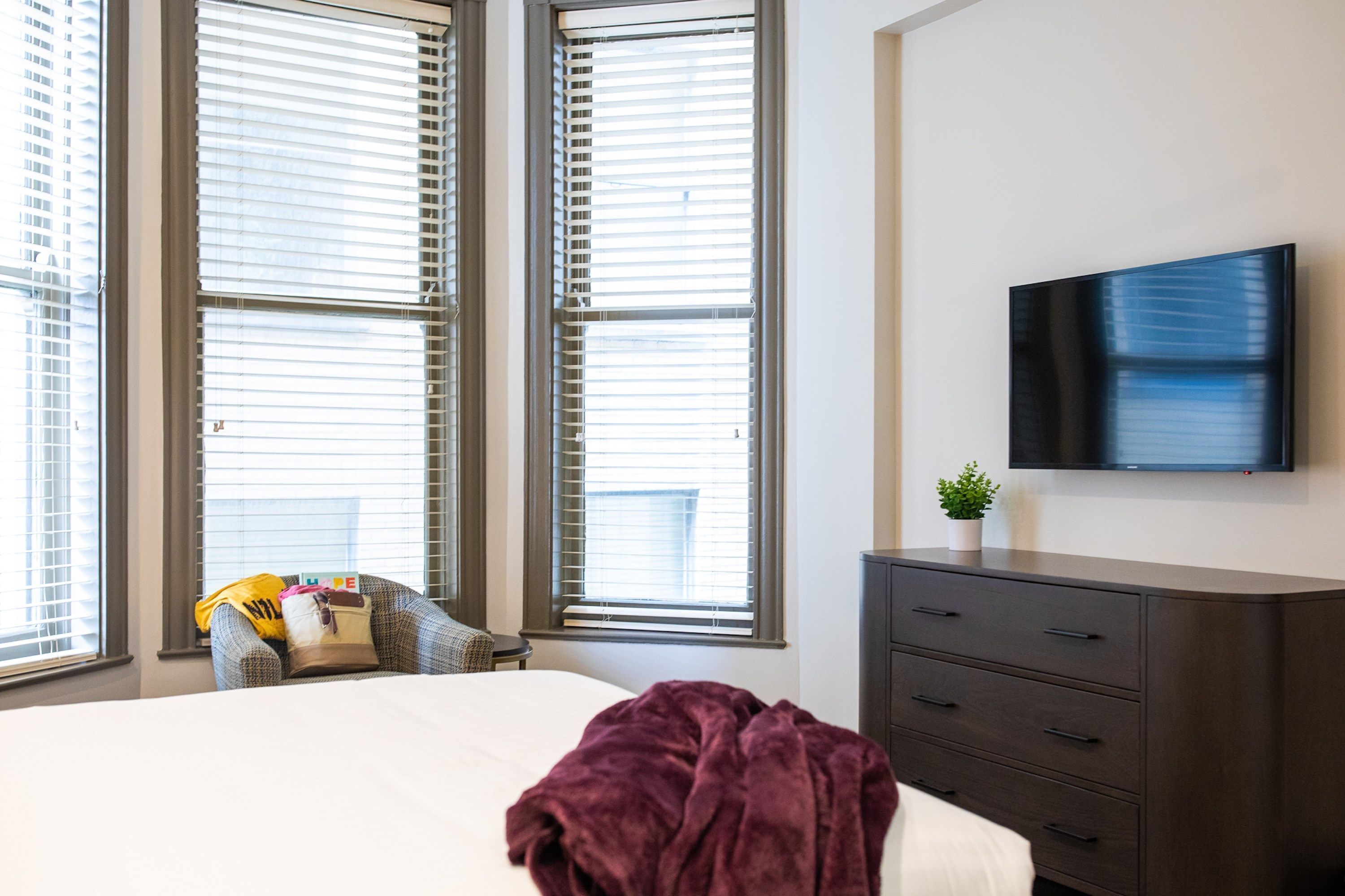 Una habitación de hotel moderna con una cama grande, un aparador de madera oscura, un televisor de pantalla plana y ventanas amplias con persianas.