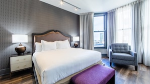 Moderni hotellihuone, jossa on suuri sänky, kaksi yöpöytää valaisimineen, tuoli, ikkuna verhoineen ja näkymä kaupunkiin.