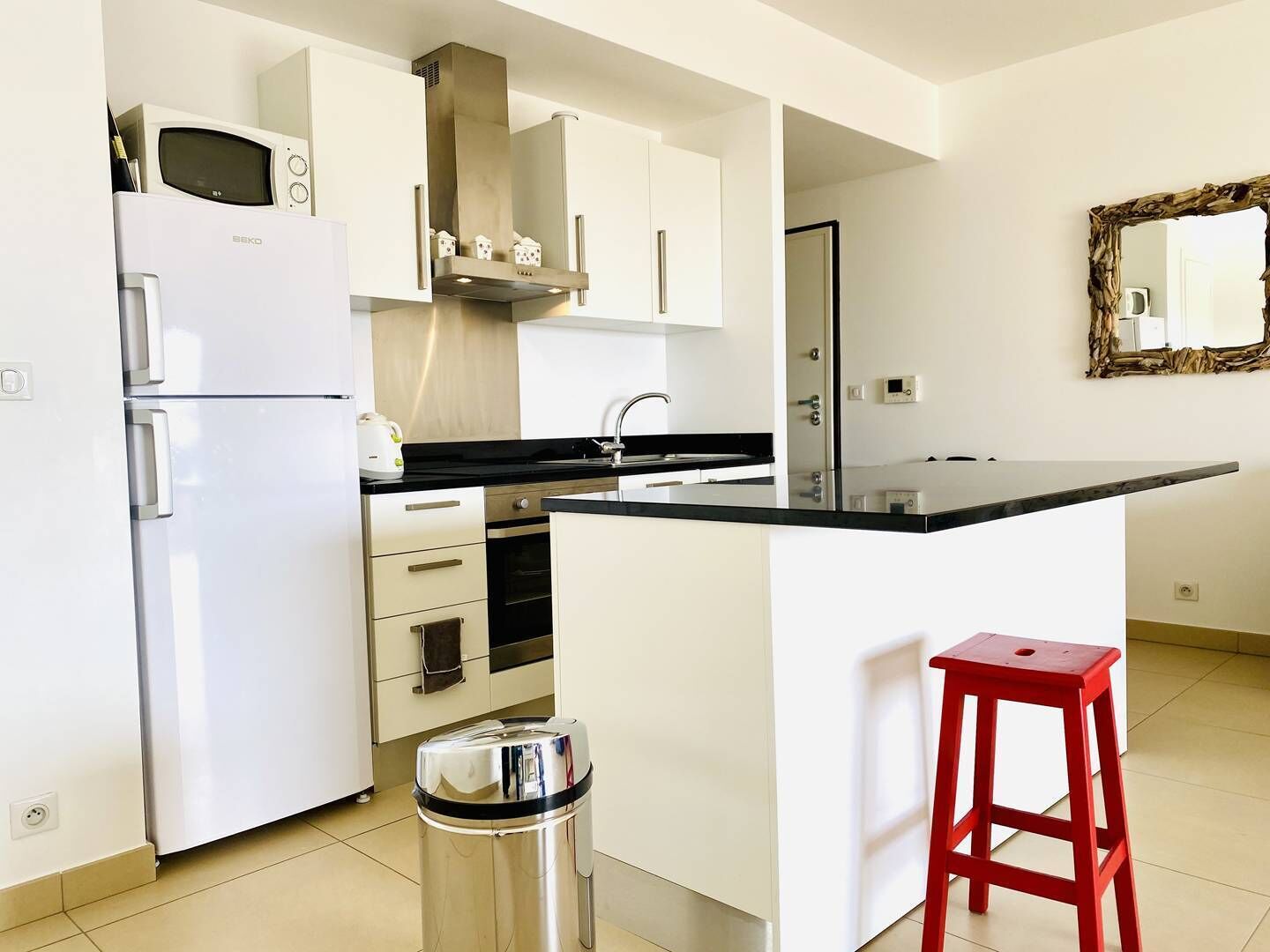 Una cocina moderna con armarios blancos, una encimera negra, un cubo de basura de acero inoxidable y un taburete rojo.