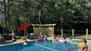 Un espace piscine où des personnes se détendent et nagent, entouré d’arbres et d’une terrasse en bois.