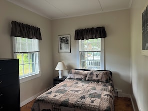 Un dormitorio con cama, dos ventanas con cortinas, una mesita de noche con lámpara, un aparador y un cuadro en la pared.