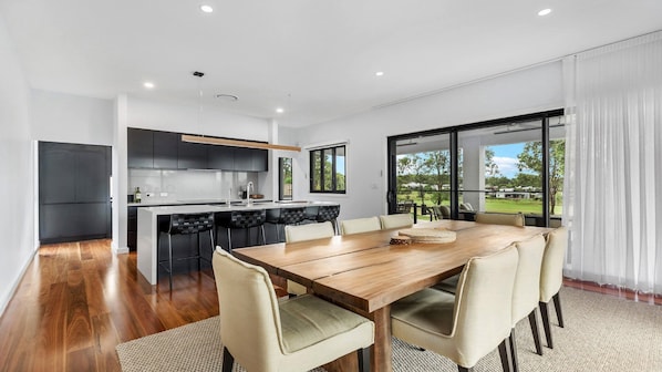 A modern kitchen with a central island, a dining area with a wooden table and chairs, and large windows offering a view of the outdoors.