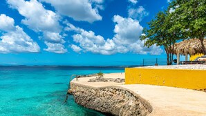Una playa con agua turquesa cristalina, un muro amarillo y una estructura con techo de paja.
