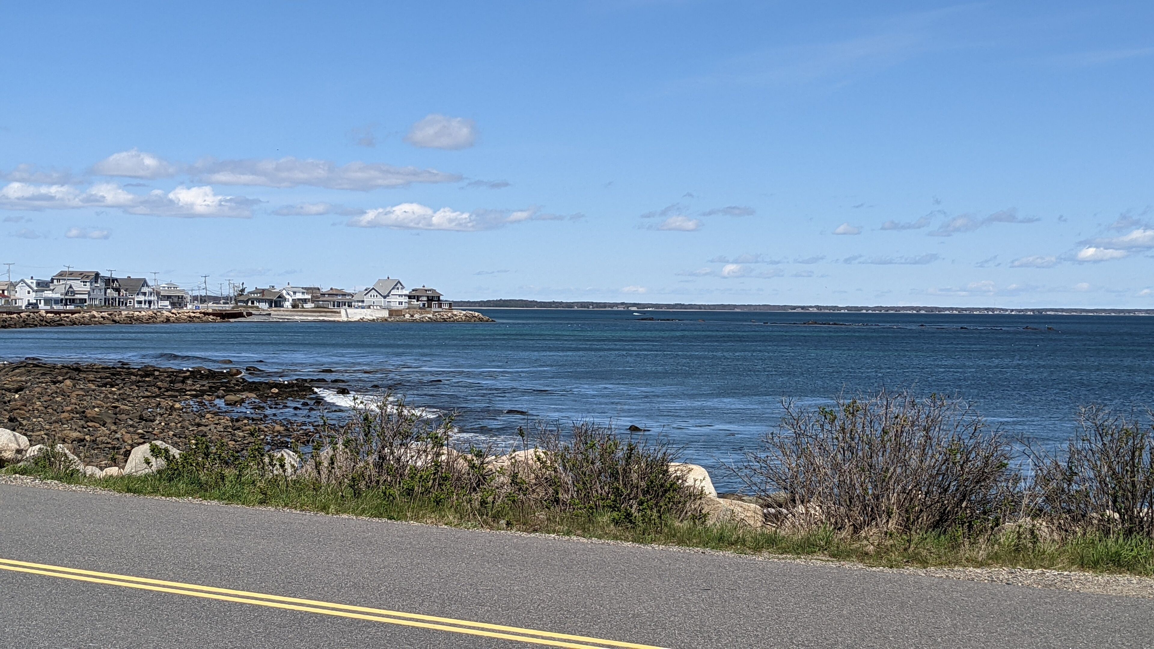 A coastal road with houses on the left and a rocky shoreline.