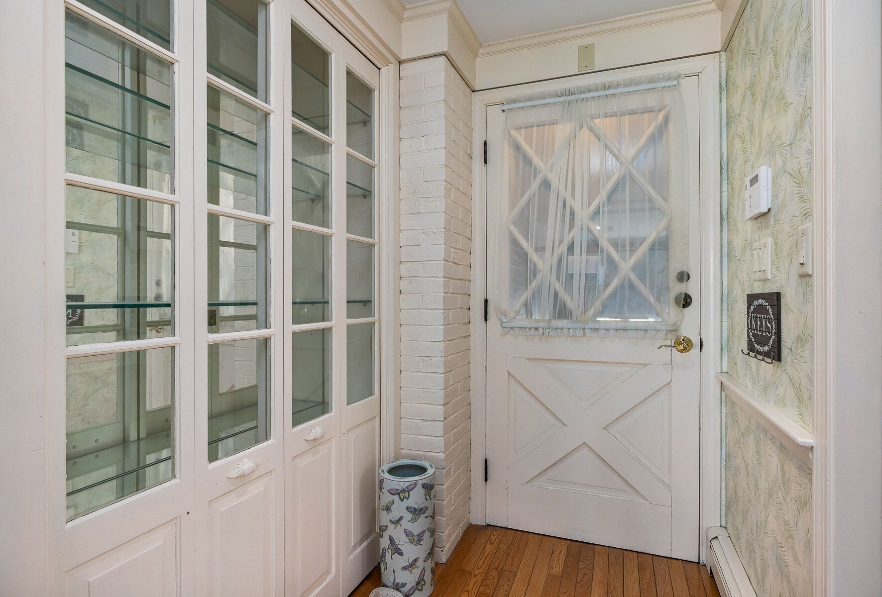 A narrow hallway with white paneled walls, a wooden floor, and a white door with a diamond pattern.