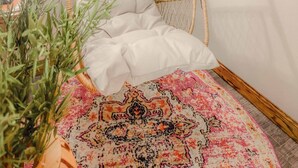 Une chaise en osier avec un coussin blanc, un tapis à motifs et une étagère avec des plantes et des haltères.