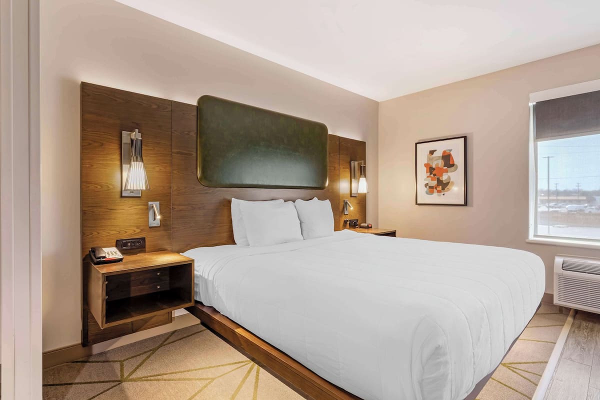Une chambre d’hôtel avec un grand lit, une tête de lit en bois, une table de chevet avec un téléphone, une lampe fixée au mur et une fenêtre donnant sur un beau paysage.