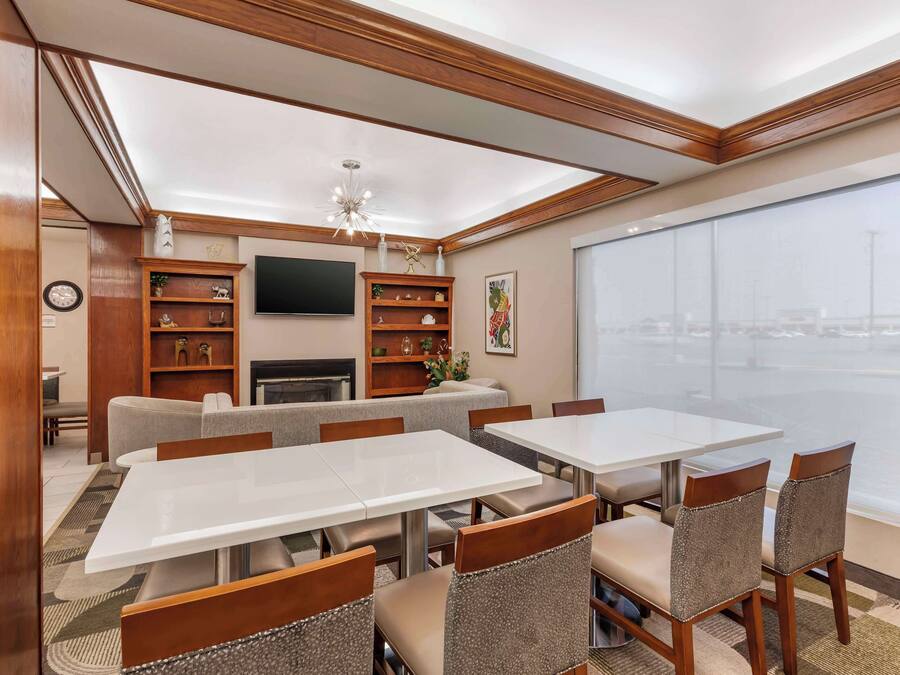 A hotel room with a dining area featuring white tables and wooden chairs, a fireplace, and a large window with a city view.