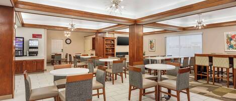 A hotel dining area with round tables and chairs, a kitchen area with a coffee machine, and a TV on the wall.