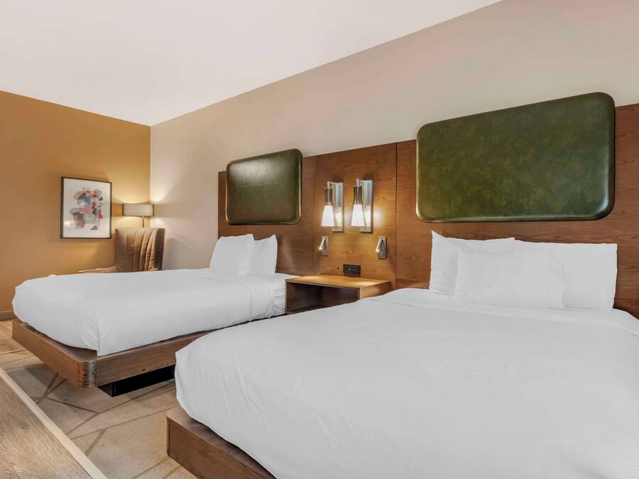 A hotel room with two single beds, wooden headboards, a nightstand with a lamp, and a framed picture on the wall.