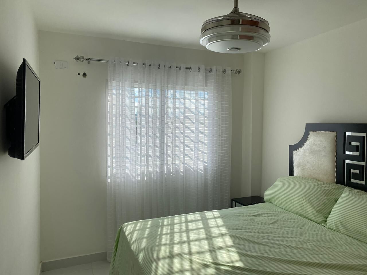 Un dormitorio con cama, una mesita de noche, un televisor montado en la pared y una ventana con cortinas translúcidas.