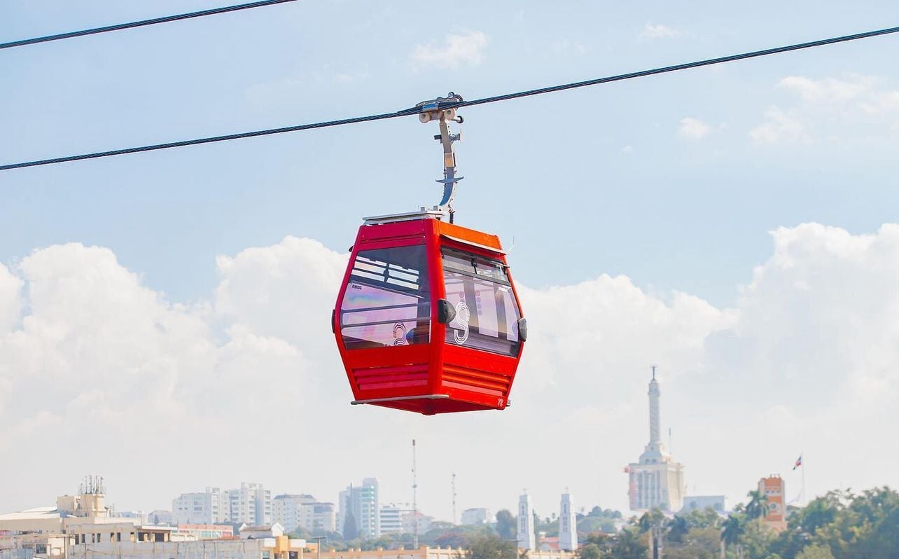 Eine rote Seilbahn, die von Stahlseilen gehalten wird, vor einer Stadtkulisse.