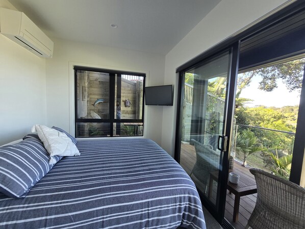A bedroom with a bed, a TV, and a balcony with a view of greenery.