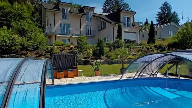 A swimming pool with a clear blue water surface, surrounded by a tiled deck. In the background, there are multi-story houses with balconies and a well-maintained garden.