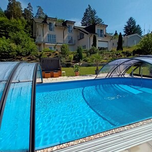 A swimming pool with a clear blue water surface, surrounded by a tiled deck. In the background, there are multi-story houses with balconies and a well-maintained garden.